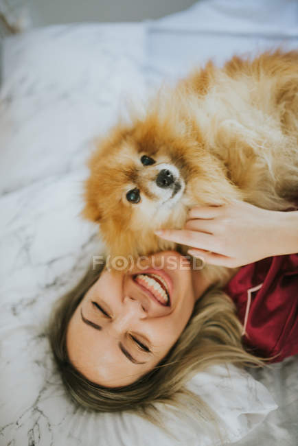 Joven feliz sonriente atractiva mujer en pijama acostado en la cama con poco peludo perro - foto de stock