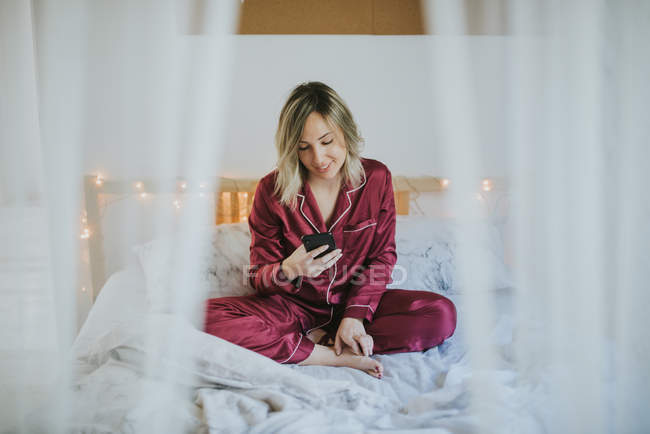 Mujer bonita joven en pijama sentada en la cama y usando un teléfono inteligente - foto de stock