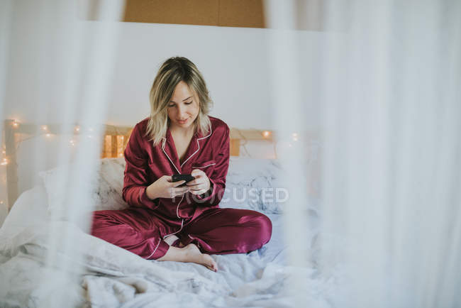 Young pretty woman in pajamas sitting on bed and using smartphone — Stock Photo