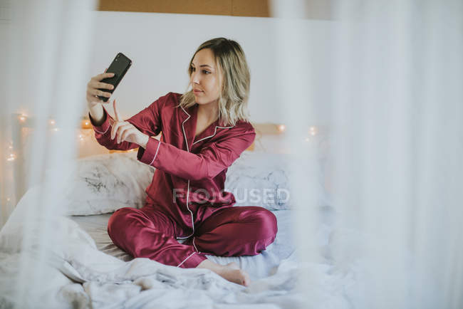 Jovem mulher bonita de pijama sentada na cama enquanto toma selfie com smartphone — Fotografia de Stock