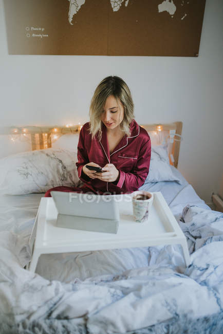 Молода симпатична жінка в піжамі за допомогою смартфона, сидячи в ліжку з кавою і планшетом на табличне підношення — стокове фото
