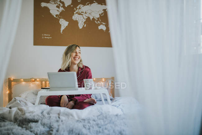 Jeune jolie femme en pyjama assise au lit avec café et tablette numérique sur plateau de table — Photo de stock