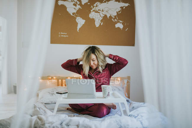 Giovane bella donna in pigiama che si estende a letto mentre seduto sul vassoio del tavolo con tablet e tazza di caffè del mattino — Foto stock