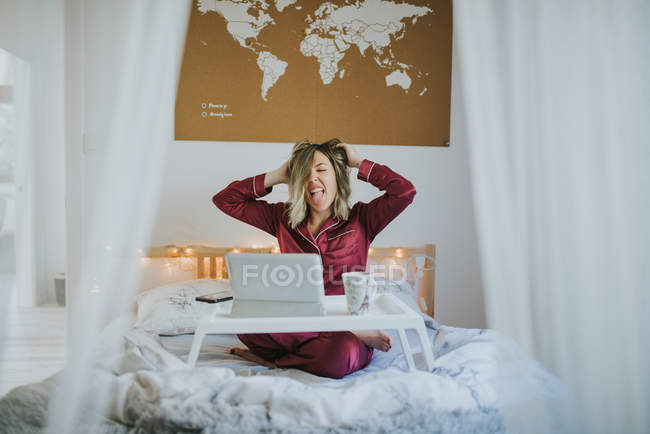 Mujer bonita joven en pijama estirándose en la cama mientras está sentado en la bandeja de la mesa con la tableta y taza de café de la mañana - foto de stock