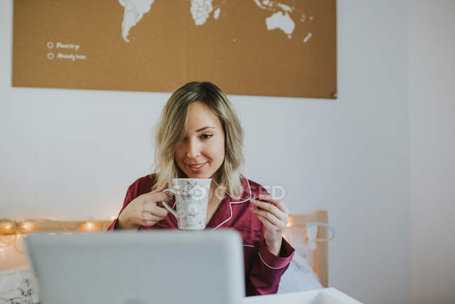 Jeune jolie femme en pyjama regardant la tablette tout en étant assis au lit avec du café — Photo de stock