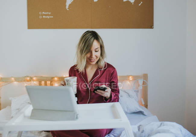Молодая красивая женщина в пижаме с помощью смартфона, сидя в постели с кофе и планшетом на подносе — стоковое фото
