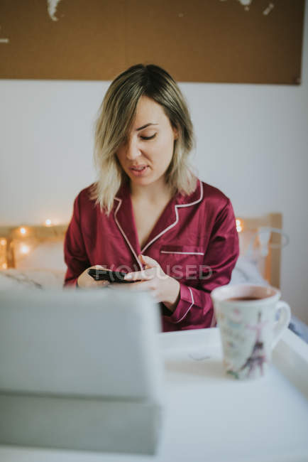 Joven mujer bonita en pijama usando teléfono inteligente mientras está sentado en la cama con café y tableta en la bandeja de la mesa - foto de stock