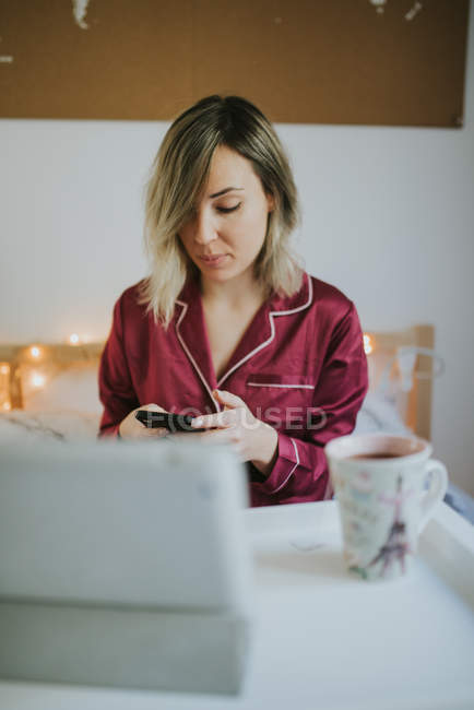 Молода красива жінка в піжамі використовує смартфон, сидячи в ліжку з кавою і планшетом на столі — стокове фото