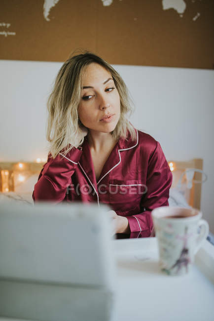 Joven mujer bonita en pijama mirando la tableta mientras está sentado en la cama con café en la bandeja de la mesa - foto de stock