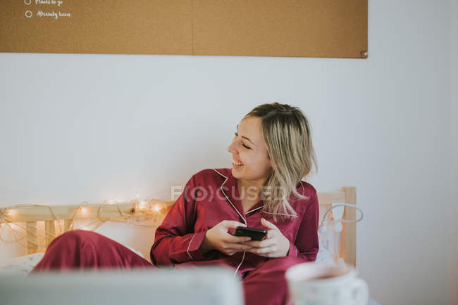 Jovem mulher rindo de pijama segurando smartphone enquanto está sentado na cama — Fotografia de Stock