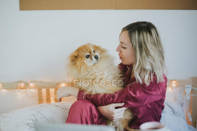 Giovane felice donna attraente sorridente in pigiama seduto a letto con piccolo cane soffice — Foto stock
