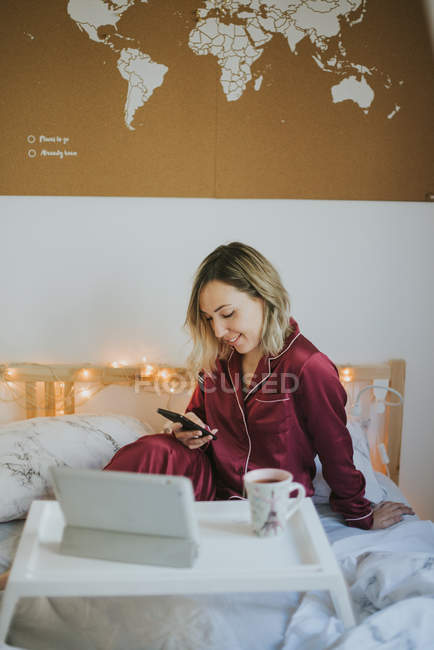 Jeune jolie femme en pyjama à l'aide d'un smartphone assis au lit avec café et tablette sur plateau de table — Photo de stock