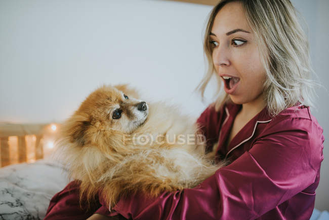 Junge glücklich lächelnde attraktive Frau im Pyjama sitzt im Bett mit kleinem flauschigen Hund — Stockfoto
