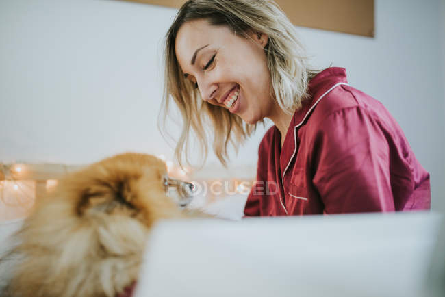 Молода щаслива усміхнена приваблива жінка в піжамі сидить у ліжку з маленькою пухнастою собакою — стокове фото