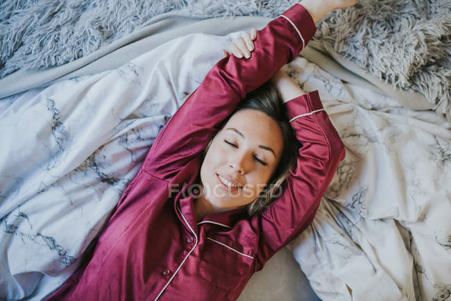 Junge hübsche Frau im Schlafanzug lächelt, während sie es sich auf dem Bett gemütlich macht — Stockfoto