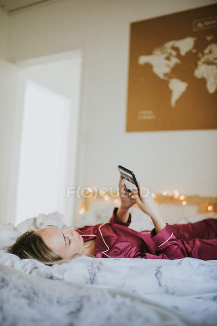 Молодая красивая женщина в пижаме лежит на кровати и использует смартфон — стоковое фото