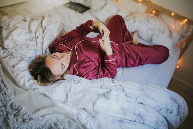 Jeune jolie femme en pyjama couchée sur le lit et utilisant un smartphone — Photo de stock