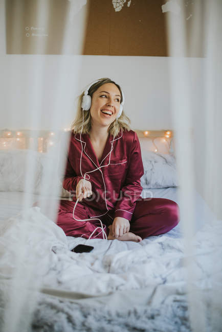 Young happy smiling woman in pajamas with headphones and smartphone sitting on bed in morning — Stock Photo