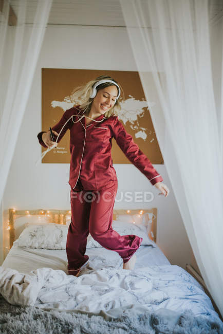 Молода щаслива усміхнена жінка в піжамі з навушниками танцює на ліжку вранці — стокове фото