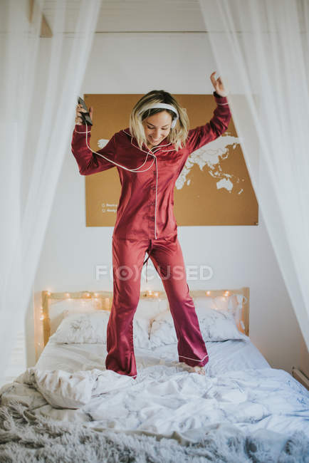 Giovane donna felice sorridente in pigiama con le cuffie che ballano sul letto al mattino — Foto stock