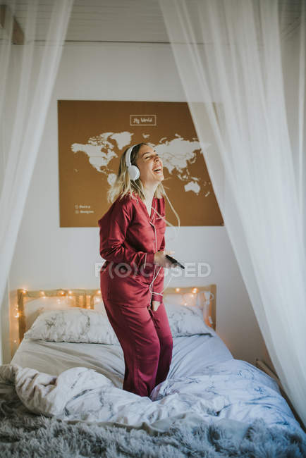 Young happy smiling woman in pajamas with headphones dancing on bed in morning — Stock Photo
