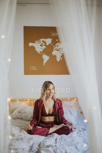 Молодая женщина в пижаме медитирует, сидя в позе лотоса на удобной кровати утром — стоковое фото