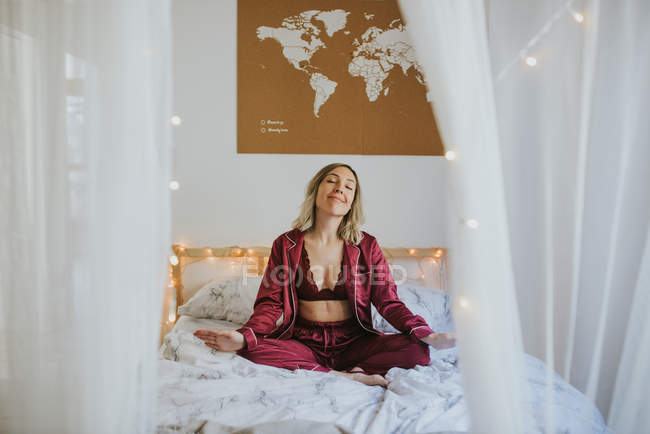 Junge Frau im Schlafanzug meditiert, während sie morgens in Lotuspose auf einem bequemen Bett sitzt — Stockfoto