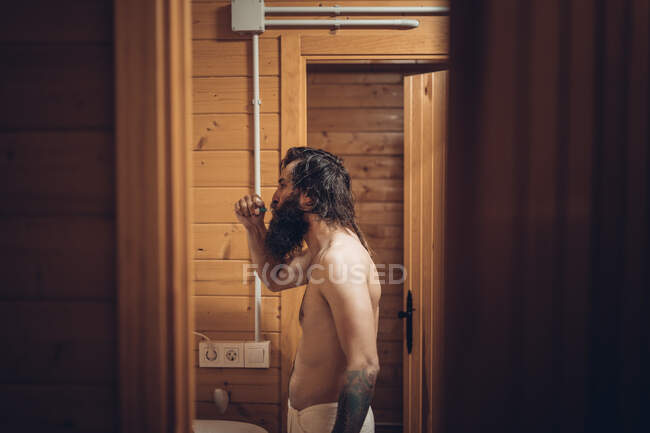 Вид збоку чоловічий хіпстер з татуюванням та чищенням бороди зубів під час відпочинку в дерев'яній кабіні — стокове фото