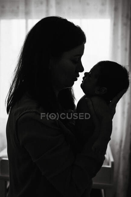 Silueta de madre joven sosteniendo las manos del bebé recién nacido mientras está de pie en casa - foto de stock