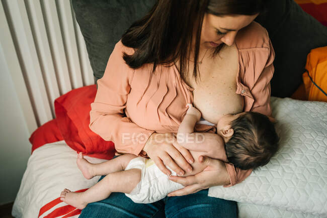 Зверху молода мати тримається за руки і годує новонародженого дитини, загорнутого в ковдру на ліжку вдома — стокове фото