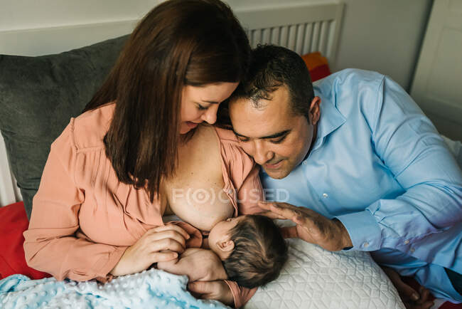 Desde arriba, madre y padre sin rostro tomados de las manos y amamantando al bebé recién nacido envuelto en una manta en la cama en casa - foto de stock