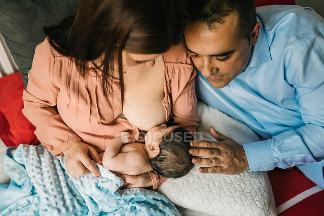 Сверху безликие мать и отец держатся за руки и кормят грудью новорожденного ребенка, завернутого в одеяло на кровати дома — стоковое фото