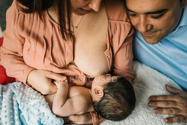 Сверху безликие мать и отец держатся за руки и кормят грудью новорожденного ребенка, завернутого в одеяло на кровати дома — стоковое фото