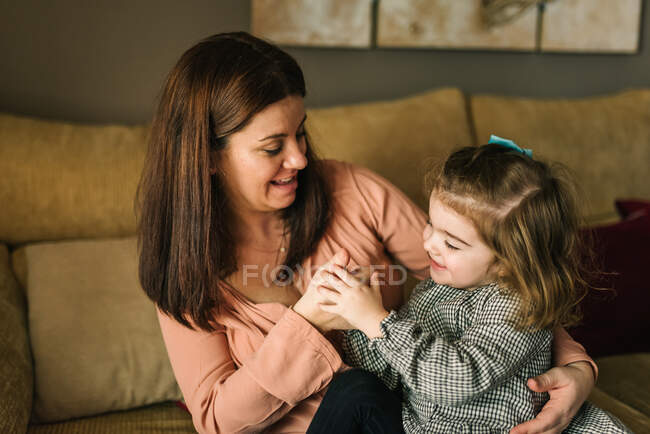 Faceless young mother embracing cheerful little daughter considering hand sitting on sofa at home — Stock Photo