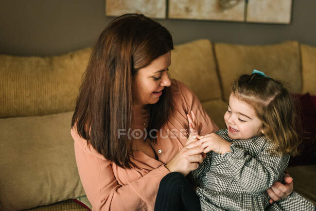 Безлика молода мати обіймає веселу маленьку доньку, розглядаючи руку, що сидить на дивані вдома — стокове фото