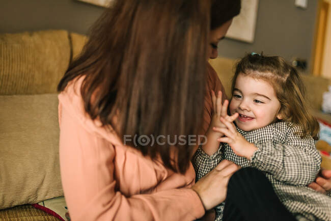 Безликая молодая мать обнимает веселую маленькую дочь, рассматривая руку, сидя на диване дома — стоковое фото