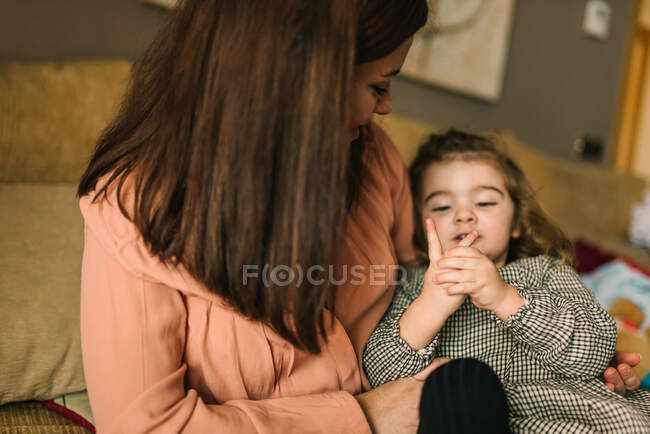 Faceless young mother embracing cheerful little daughter considering hand sitting on sofa at home — Stock Photo