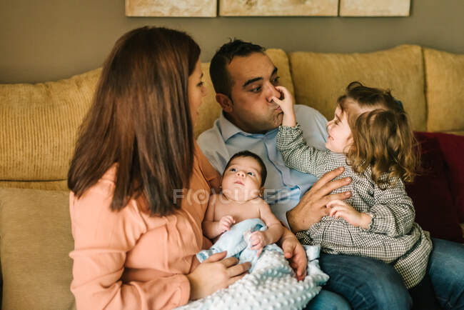 Щаслива молода мати тримає новонароджену дитину, загорнуту в ковдру, а батько тримає дочку, сидячи на дивані вдома — стокове фото