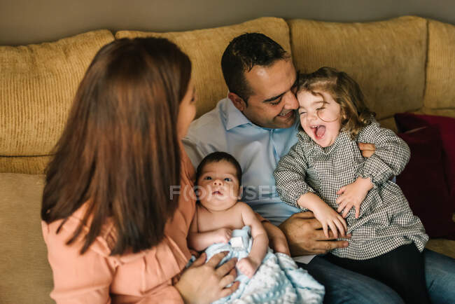 Joyeux jeune mère tenant le nouveau-né enveloppé dans une couverture et père tenant sa fille assise sur le canapé à la maison — Photo de stock