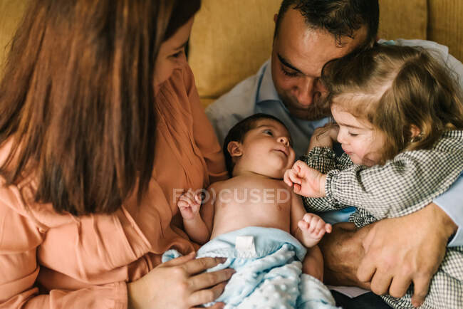 Happy young mother holding newborn baby wrapped in blanket and father holding daughter sitting on sofa at home — Stock Photo