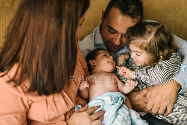 Счастливая молодая мать держит новорожденного ребенка, завернутого в одеяло, а отец держит дочь, сидящую дома на диване — стоковое фото