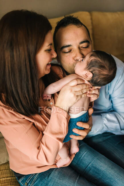 Glückliche junge Mutter hält Neugeborenes in Decke gewickelt und Vater küsst Baby auf Sofa zu Hause — Stockfoto