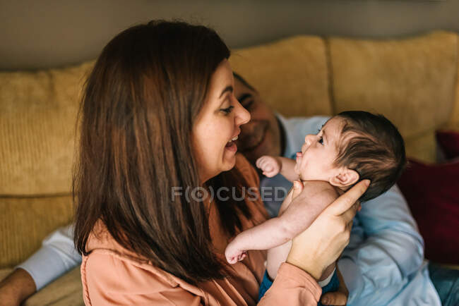 Счастливая молодая мать держит новорожденного ребенка, завернутого в одеяло, а отец сидит на диване рядом с ними дома — стоковое фото