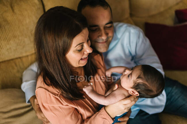 Feliz madre joven sosteniendo al bebé recién nacido envuelto en manta y padre sentado en el sofá junto a ellos en casa - foto de stock