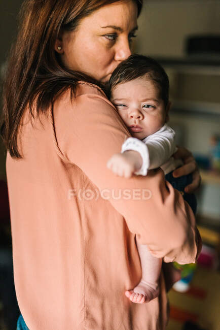 Volver ver morena mamá abrazando lindo bebé mirando hacia fuera en casa - foto de stock