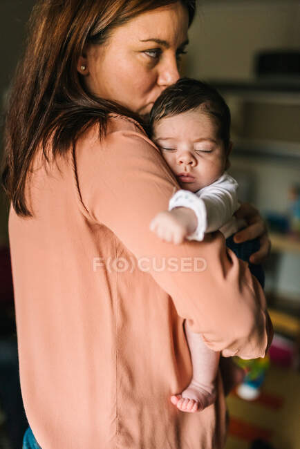 Volver ver morena mamá abrazando lindo bebé mirando hacia fuera en casa - foto de stock
