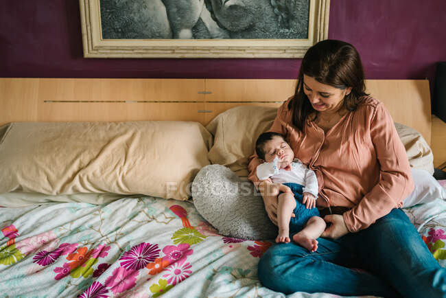 Joven madre abrazando alegre pequeña hija sentada en la cama en casa - foto de stock