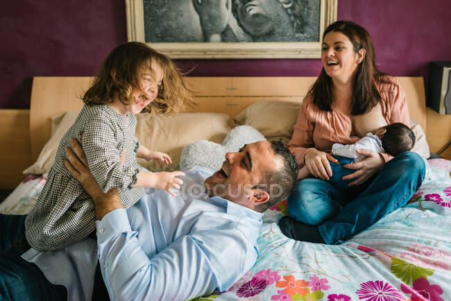Smiling father holding laughing little daughter lying on bed with mother breastfeeding newborn baby on background in bedroom — Stock Photo