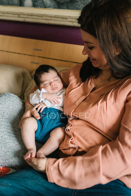 Молодая мать обнимает веселую маленькую дочь сидя на кровати дома — стоковое фото