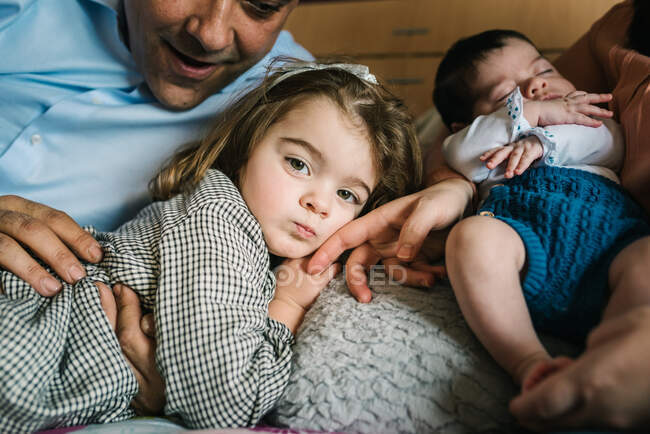 Sonriente padre sosteniendo riendo riendo pequeña hija acostada en la cama con la madre sosteniendo al bebé recién nacido en el fondo en el dormitorio - foto de stock